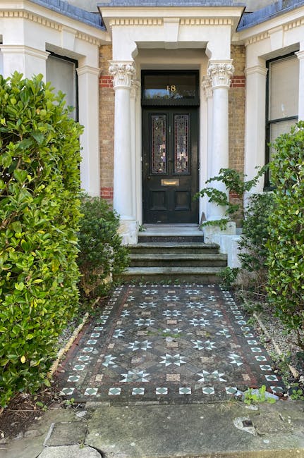 A large, three-storey red brick building with a symmetrical façade, featuring decorative window mouldings and a central arched doorway with a black wooden door, situated in a landscaped area with tri