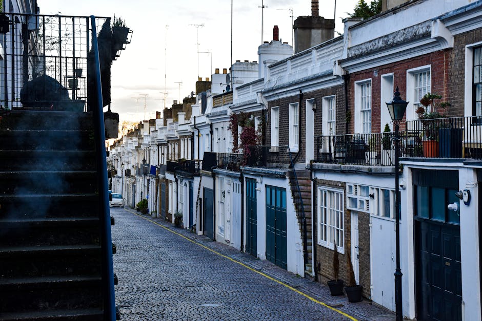 A narrow street in a residential area with a sloped, cobblestone pavement lined by row houses featuring brick and painted facades, some with small balconies and potted plants. On the left, a black met